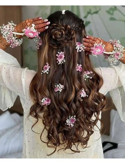 A beautiful Mehendi hairstyle with a rose-shaped bun and flowing curls. The hair is decorated with fresh pink floral accessories to match the festive theme.