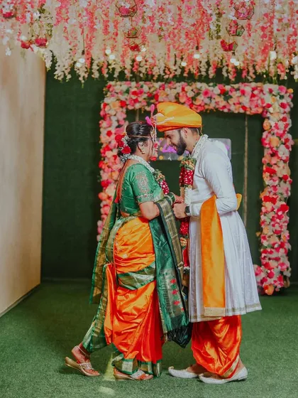 A beautiful portrait of the couple facing each other, framed by a floral arch. The bride is wearing a stunning orange and green Nauvari saree, showcasing traditional Maharashtrian bridal wear.