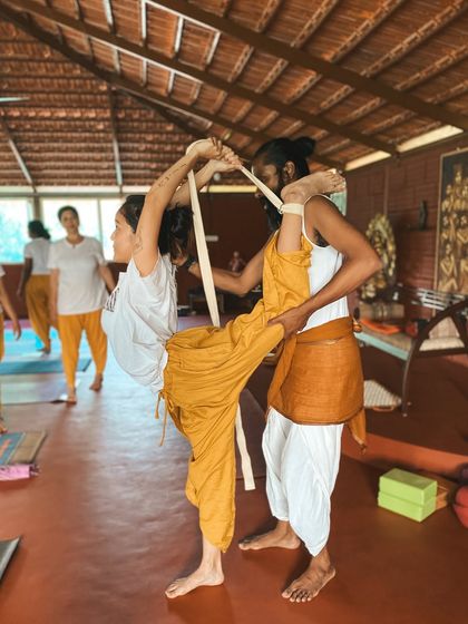 With the help of a strap, I assist a student in an advanced standing split variation. This demonstrates how we can work towards deeper flexibility with safety and support, honoring the body's current abilities while gently encouraging growth.