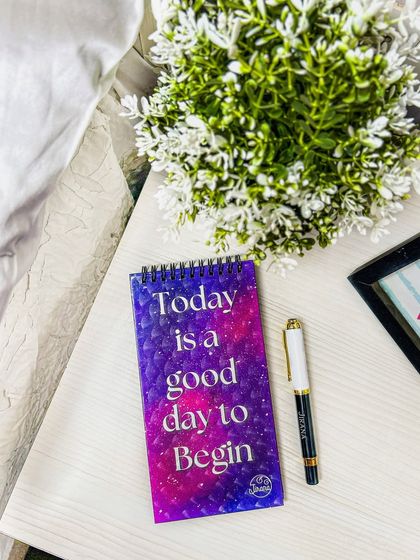 A close-up of a 'Today is a good day to Begin' notepad, styled in a cozy bedroom setting. This lifestyle shot creates an inspirational and calming mood.