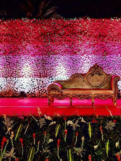 An outdoor reception setting under a canopy of fairy lights. The stage is bathed in a warm golden glow, and the guest tables are decorated with pink accents.