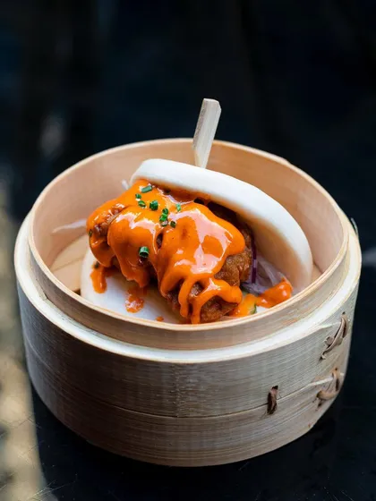 A fluffy bao bun filled with crispy fried chicken and a vibrant orange sauce, served in a traditional bamboo steamer. The presentation is as appealing as the food itself.