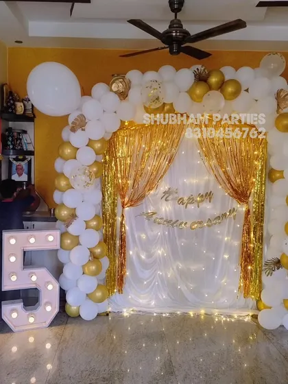 Celebrate a milestone 25th anniversary with this elegant and grand decoration. The setup includes a beautiful white and gold balloon arch, a shimmering backdrop with fairy lights, and large light-up marquee numbers to mark the special occasion.