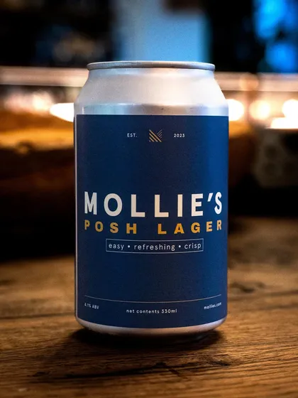 A can of Mollie's Posh Lager in a cozy, candlelit setting. The ambiance makes the product feel like part of a relaxed, premium experience.