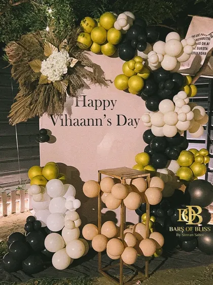 A simple yet stylish birthday backdrop for 'Vihaann's Day'. The decor uses a neutral color palette of beige, black, and white balloons, accented with pampas grass for a modern and understated look.