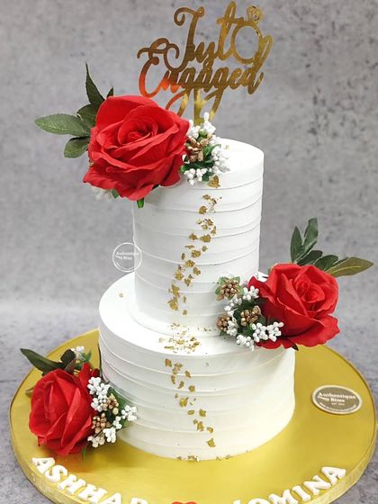 A bold and beautiful two-tiered engagement cake. The crisp white tiers are adorned with vibrant red roses and subtle gold leaf, creating a striking and passionate design for a "Just Engaged" celebration.