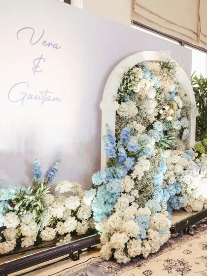 A chic photo backdrop with the couple's names, featuring a floral installation bursting from an arched window frame, blending modern design with classic floral beauty.