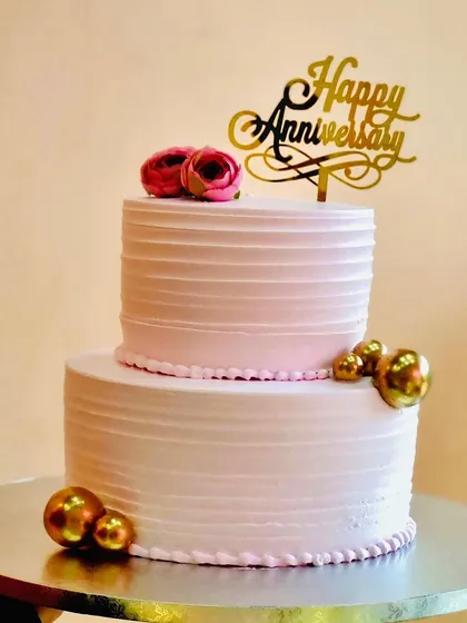 Simple elegance defines this two-tier anniversary cake. The soft pink frosting with clean lines, accented by gold spheres and a "Happy Anniversary" topper, creates a sophisticated look for celebrating another year together.