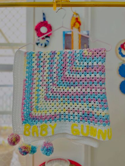 This pastel granny square blanket, customized with "Baby Gunnu," is a classic choice. The addition of multi-colored pom-poms on the corners makes it extra special and fun for a little one.