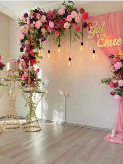 A stunning floral arch for an engagement or anniversary. The setup features lush arrangements of pink and red roses, elegant draping, and vintage-style Edison bulbs for romantic lighting.