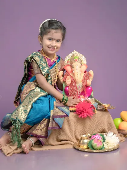 For the Ganpati festival, I dress your child in beautiful traditional Maharashtrian attire, like this girl in a Paithani-style saree. The setup includes a Ganpati idol, a puja thali, and fresh fruits to create an authentic festive atmosphere.