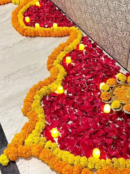 A traditional floor decoration (rangoli) made with flower petals for a housewarming event. This design uses vibrant yellow marigolds and red rose petals, outlined with small electric candles for a warm glow.