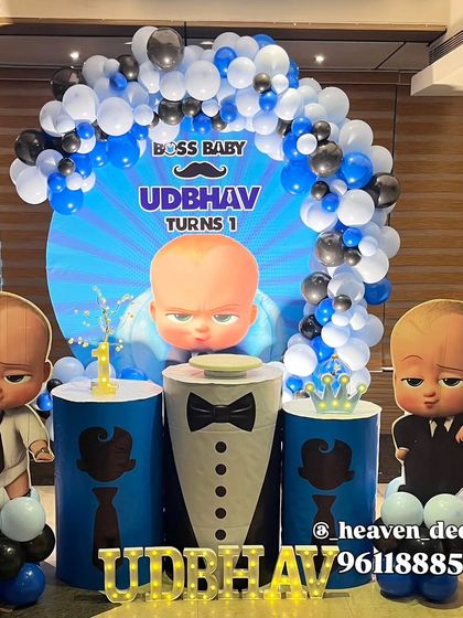 A dynamic Boss Baby first birthday with a circular backdrop featuring a sunburst design. The setup includes character cutouts, themed plinths, and a balloon arch in blue, black, and white.