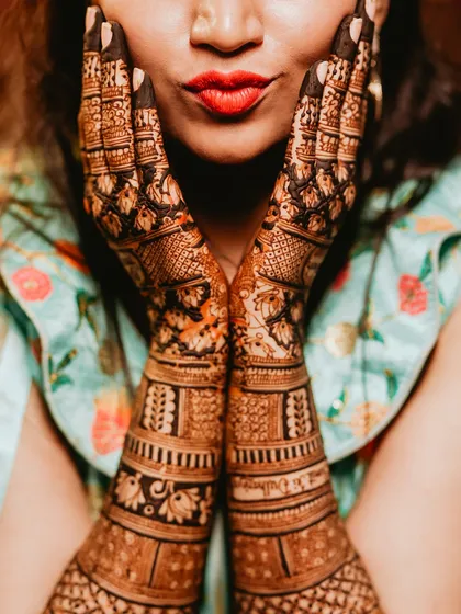 A close-up shot framing the bride's face with her henna-covered hands. The vibrant red lipstick provides a beautiful pop of color.