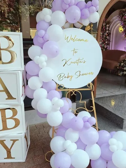 A classic baby shower welcome area. The setup includes "BABY" blocks and a welcome sign on an easel, both decorated with a soft lavender and white balloon garland.