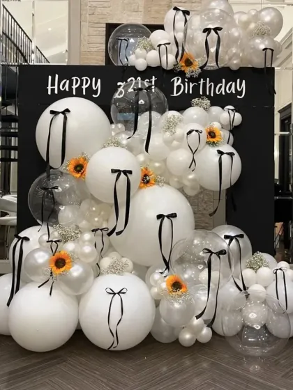 A chic and modern 21st birthday backdrop in black and white. The design uses large white balloons tied with black ribbons and pops of sunflower yellow for a unique, high-fashion look.