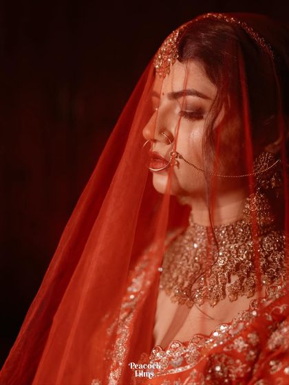 A close-up bridal portrait, with the sheer red veil creating a soft, dreamy effect. This image captures the bride's delicate features and the rich color of her wedding lehenga.