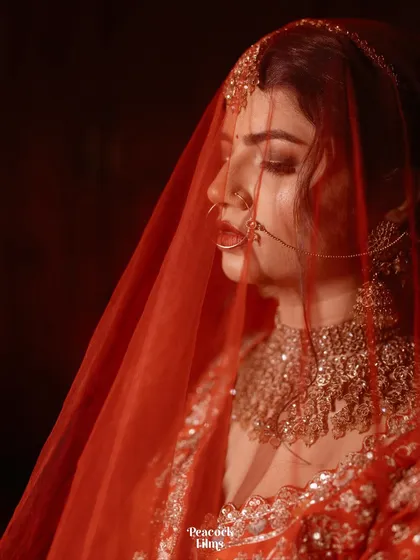 A close-up bridal portrait, with the sheer red veil creating a soft, dreamy effect. This image captures the bride's delicate features and the rich color of her wedding lehenga.