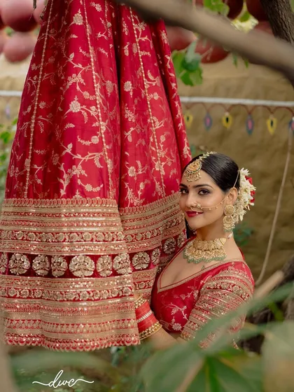 A beautiful bridal portrait, with the bride peeking out from behind her stunning red wedding lehenga, her smile full of anticipation.