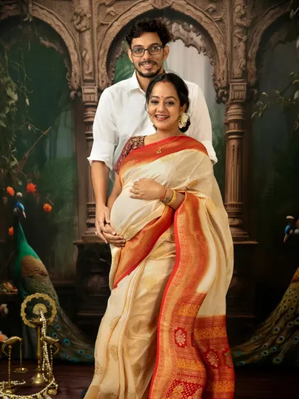 A stunning portrait of a couple expecting their baby, posing in front of a regal, peacock-themed backdrop that complements the mother's traditional saree.