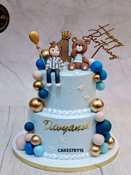A royal teddy bear celebration cake for a first birthday. This 3kg two-tier chocolate cake features a fondant boy and teddy topper, decorated in a handsome blue and gold color scheme.