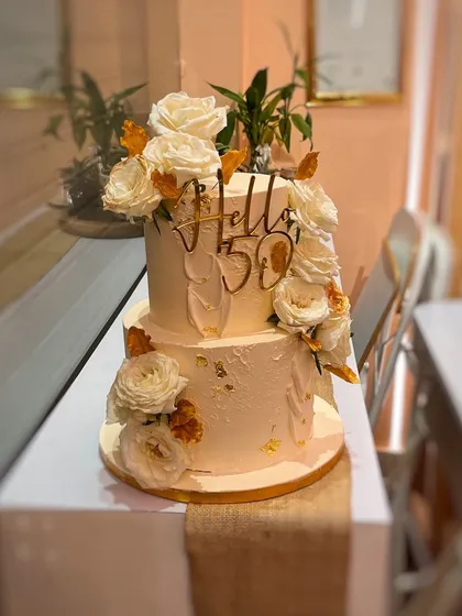 "Hello 50" has never looked so good. This two-tier cake features a textured buttercream finish, adorned with an abundance of fresh white roses and gold leaf details, making it perfect for a milestone celebration or an elegant wedding.