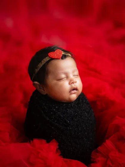 A bold and beautiful setup with a newborn girl wrapped in black against a vibrant red, textured backdrop. The tiny heart headband adds the perfect sweet detail.