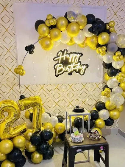 A 27th birthday party with a circular balloon arch in black and gold. The setup includes a "Happy Birthday" neon sign and foil number balloons.