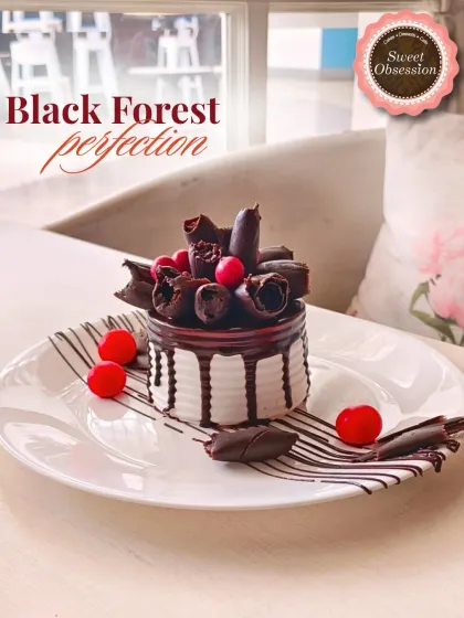 A classic Black Forest pastry, reimagined. This individual portion features layers of chocolate sponge, cream, and cherries, beautifully decorated with chocolate curls.