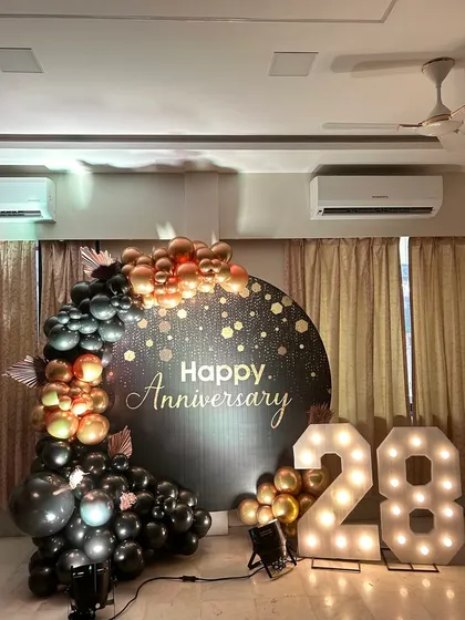 A chic 28th anniversary decoration for a home celebration. This setup features a stylish black backdrop with gold confetti details, a black and gold balloon garland, and large light-up numbers.