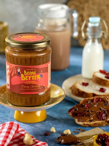 A styled product shot of Being Better's chocolate peanut spread. The presentation on a cake stand with bread and ingredients in the background shows the product in a delicious, ready-to-eat context.