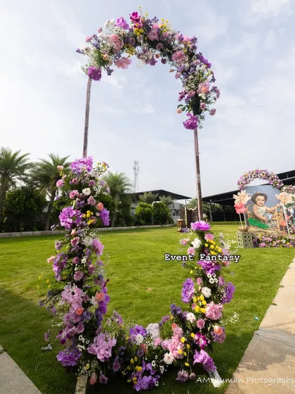 A beautiful floral horseshoe arch designed for an outdoor naming ceremony. The abundance of purple and pink flowers creates a stunning photo opportunity on the green lawn.