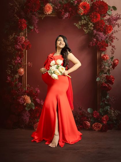A classic fine art maternity pose. The mother-to-be stands elegantly in a vibrant red gown, framed by a beautiful arch of red flowers that complements her dress perfectly.
