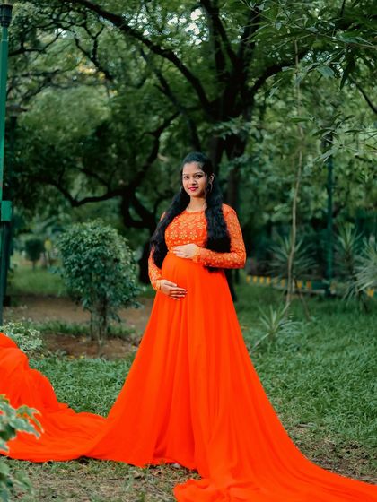 A magnificent outdoor shot featuring a mother-to-be in a vibrant orange gown with a long, flowing train. The lush green park provides a beautiful natural backdrop.