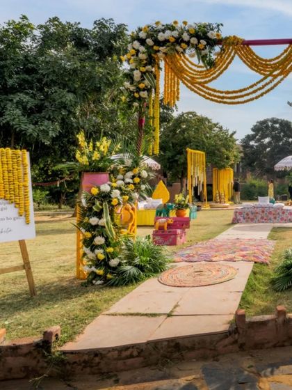 An inviting entrance to an outdoor Haldi ceremony. The path is lined with colorful decor elements, leading towards the main event area decorated with marigold garlands.