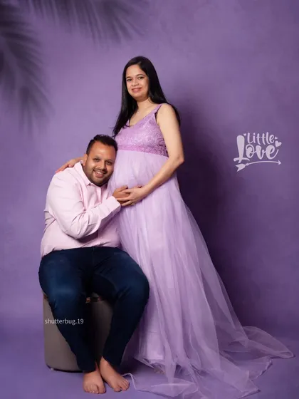 A happy couple's portrait against a soft purple backdrop. The mom-to-be is standing in her flowing gown while her partner sits, creating a sweet and connected pose.