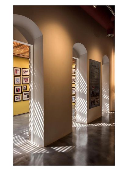 A gallery corridor where a rough, exposed stone wall of the original structure is juxtaposed with a smooth, yellow exhibition wall. Light filtering through the trellised ceiling creates rhythmic patterns, adding another layer of texture to the space.