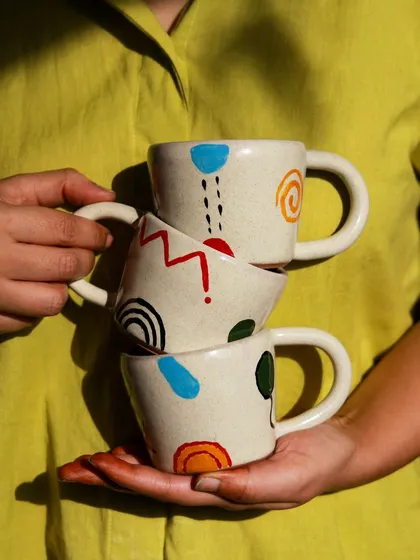 A stack of three mugs from the 'Playful Mornings' collection, each with a unique, hand painted design, held in hand to show their playful character.