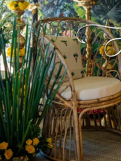 A cozy rattan chair set against a tropical print backdrop. The combination of natural textures, lush greenery, and marigold flowers creates an earthy and lively atmosphere for a Haldi celebration.