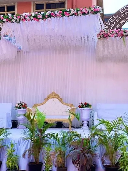 A clean and classic all-white stage setup for a daytime event. The simplicity of the white drapes and floral tassels is complemented by a row of green plants for a touch of natural color.