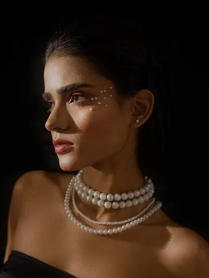 A classic profile beauty shot against a dark background. The lighting carves out the model's features and highlights the layered pearl necklace and eye makeup.