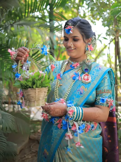 A lovely look for a baby shower, featuring a sky-blue saree and custom floral jewelry in pink and blue. The makeup is fresh and bright, and you can see the special 'आई' brooch pinned to her saree.