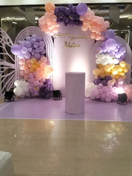 A stunning butterfly-themed first birthday stage in shades of lavender and peach. The decor features large butterfly wing cutouts, a light-up number '1', and beautiful balloon cascades, creating a dreamy and elegant backdrop.