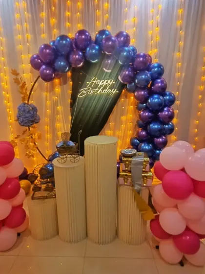 A stylish birthday decoration with a deep blue and pink balloon arch. A dark green drape and "Happy Birthday" neon sign create a unique backdrop.