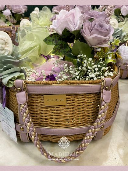 A beautifully crafted cane arrival basket designed for a baby girl's announcement. The basket is filled with lavender and white flowers and features elegant details like a cameo-style angel motif and braided lavender leather straps.