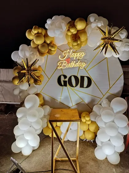 A stylish birthday decoration with a white and gold theme. The setup includes a custom geometric-patterned backdrop and a balloon arrangement accented with star-shaped foil balloons.