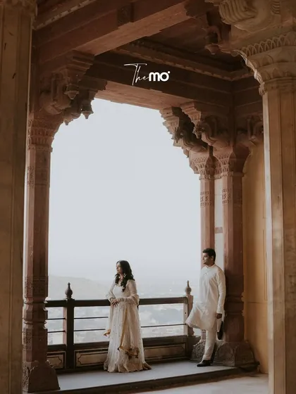 A contemplative portrait of a couple looking out from a palace balcony in Jaipur. The grand archway frames them against the sky, creating a sense of scale and quiet grandeur.