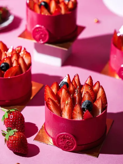 A row of elegant individual strawberry mousse cakes with a vibrant red glaze. The hard light and shadows create a modern, graphic look that emphasizes the desserts' perfect form.