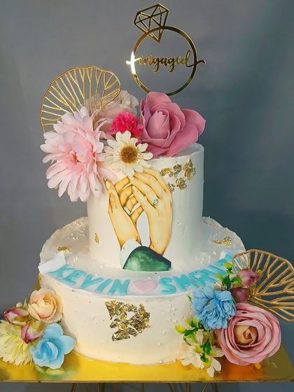 A two-tiered engagement cake featuring an illustration of holding hands, surrounded by fresh flowers and golden fan toppers.