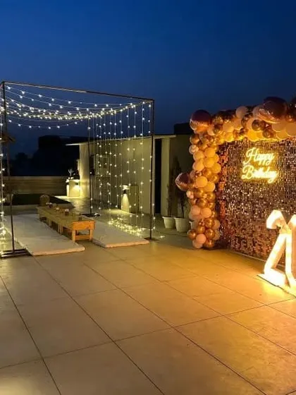 A stunning terrace party setup for a 40th birthday. The design features a large cabana frame draped with fairy lights for evening celebrations, a golden sequin backdrop with a neon sign, and a balloon arch.